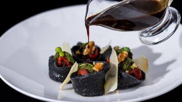 Un plato blanco con un alimento de color oscuro cubierto de nueces y hojas verdes, y una salsa oscura siendo servida sobre él.