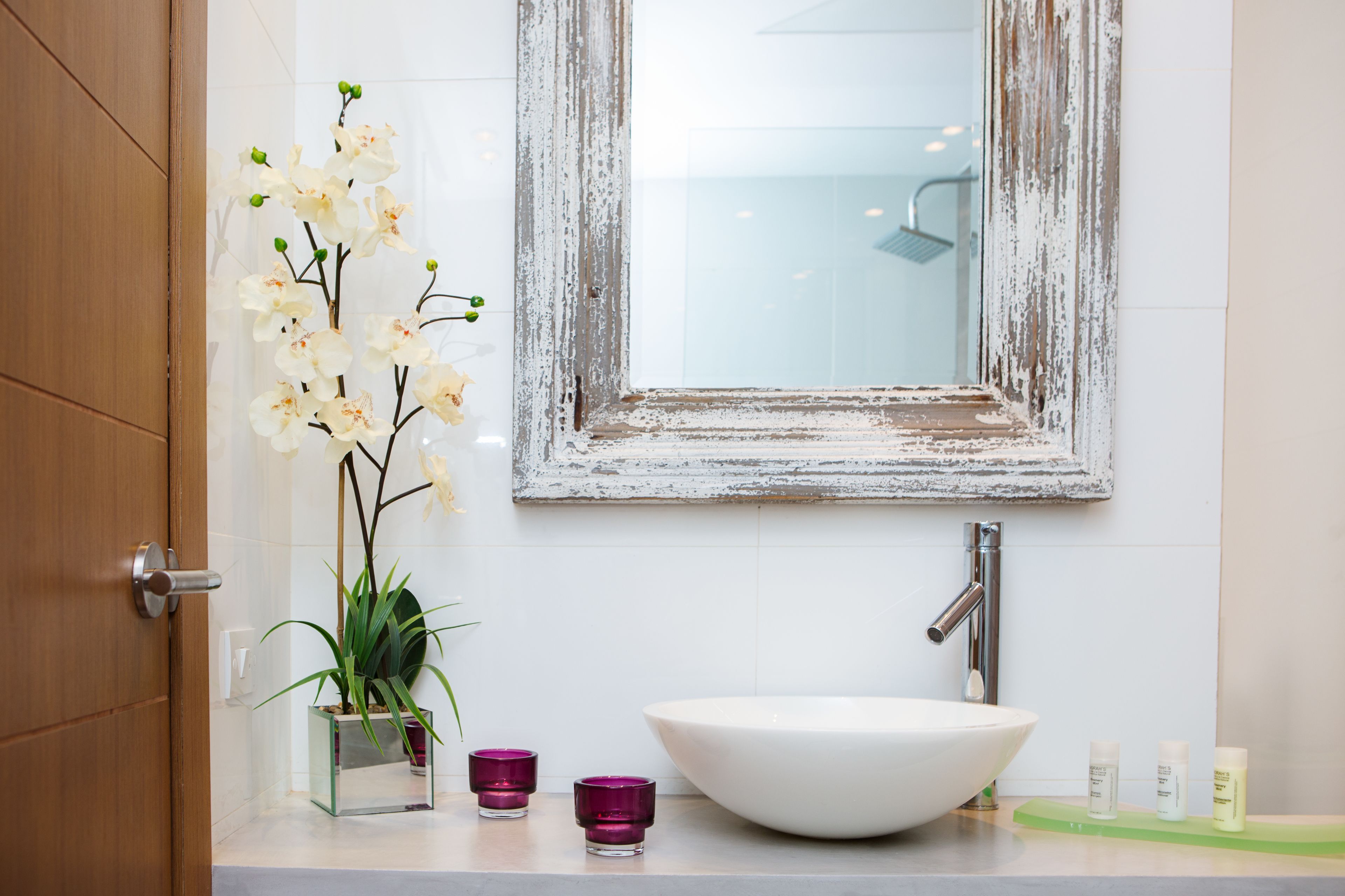 Ein Badezimmer mit einem weißen Waschbecken, einem Spiegel mit verwitterter Rahmung, einer Topf-Orchidee und zwei violetten Kerzen.