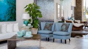 Una sala moderna con un sofá blanco, un sillón azul y una mesa de centro. Hay una planta grande y un tapiz decorativo.