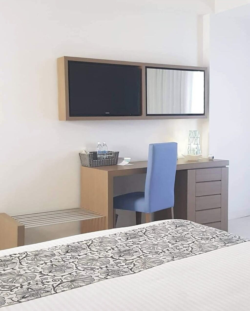 Une chambre d’hôtel comprenant un lit, un bureau avec une chaise, un téléviseur à écran plat, une fenêtre avec des rideaux, ainsi qu’une table de chevet avec une lampe et un panier.