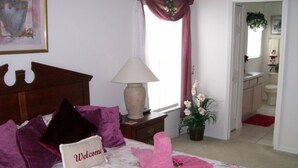 A bedroom with a bed, a bedside table with a lamp, a welcome sign, and a window with curtains. - 4BR 3BA Home in Indian Creek by CV-2653 (Kissimmee)
