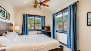 Quarto com cama, ventoinha no teto, televisão e amplas janelas com cortinas.