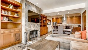 A modern living room with a fireplace, wooden shelving, and a dining area with a kitchen in the background. - Ski in, Ski out, 2 Bedroom Luxury Residence with hot tub access and expansive views (Snowmass Village)