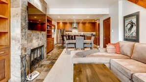 A modern living room with a fireplace, a dining area, and a kitchen with wooden cabinets and a stone backsplash. - Ski in, Ski out, 2 Bedroom Luxury Residence with hot tub access and expansive views (Snowmass Village)