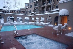 A heated outdoor pool area with lounge chairs and a hot tub, surrounded by buildings. - Snowmass Woodrun 9956 (Snowmass Village)