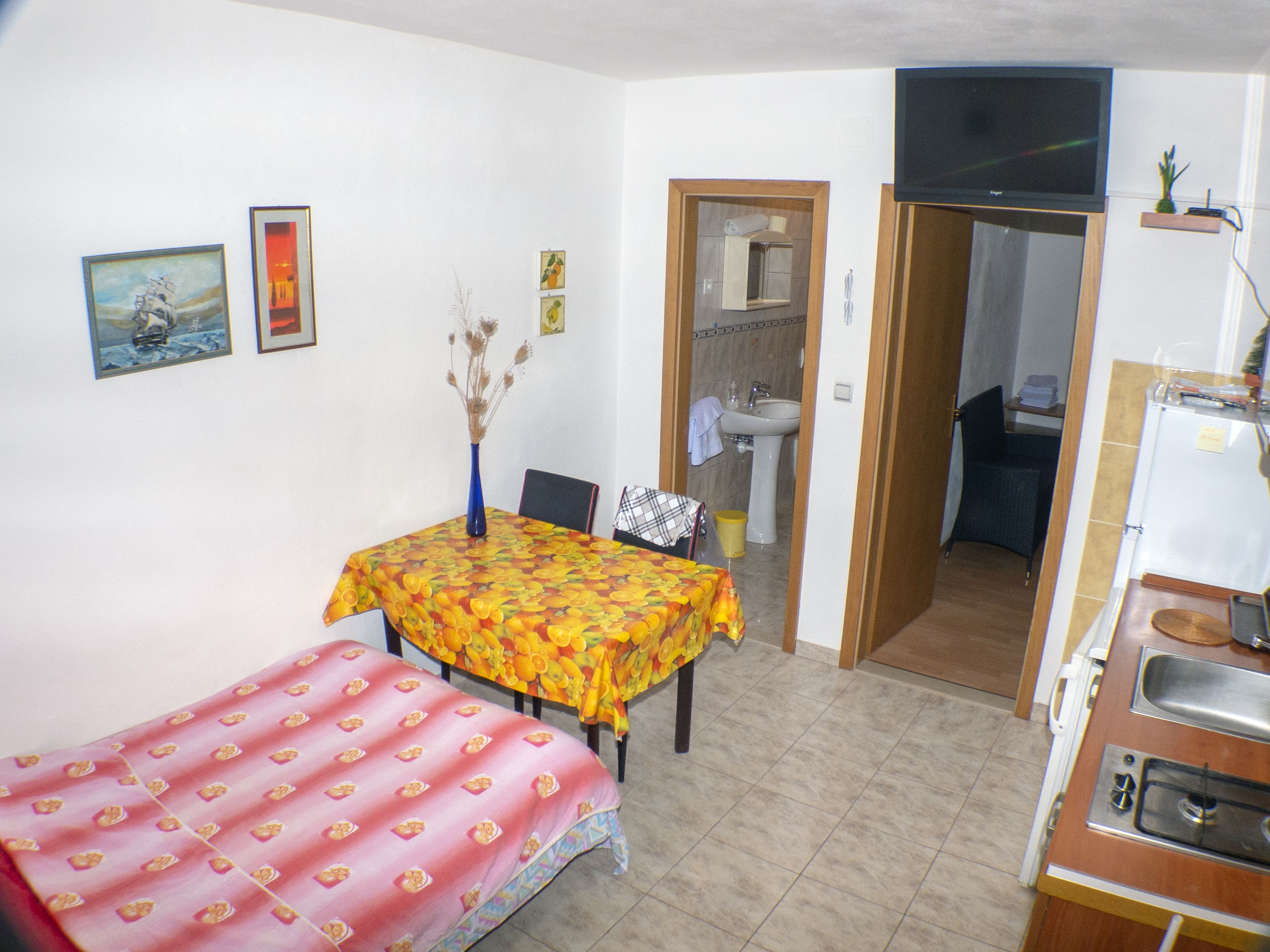 Una habitación pequeña y funcional con cama, una mesa con mantel estampado de frutas, televisor montado en la pared y un rincón de cocina.