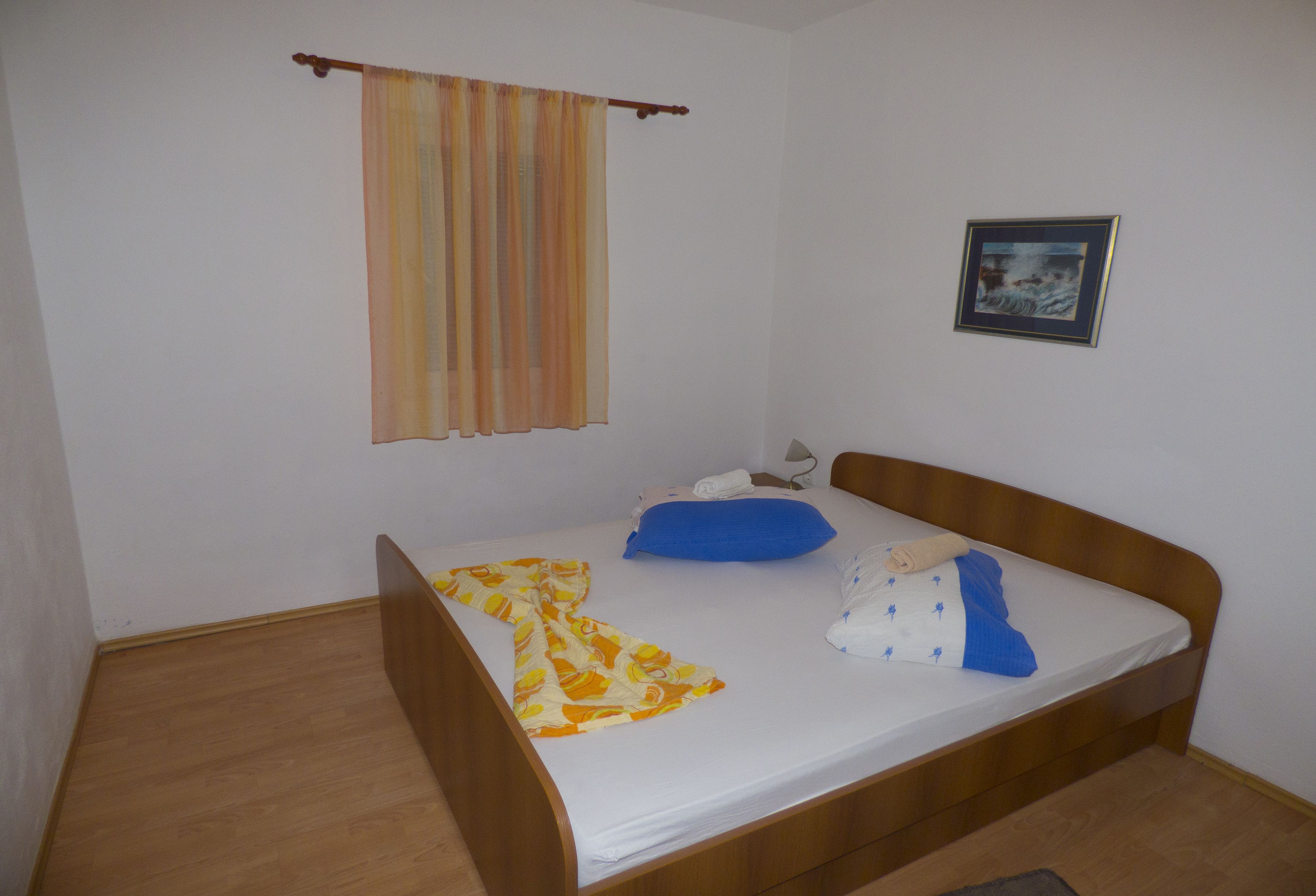 A single bed with a wooden frame, a blue pillow, a white pillow with a blue cover, and a yellow and orange striped curtain.