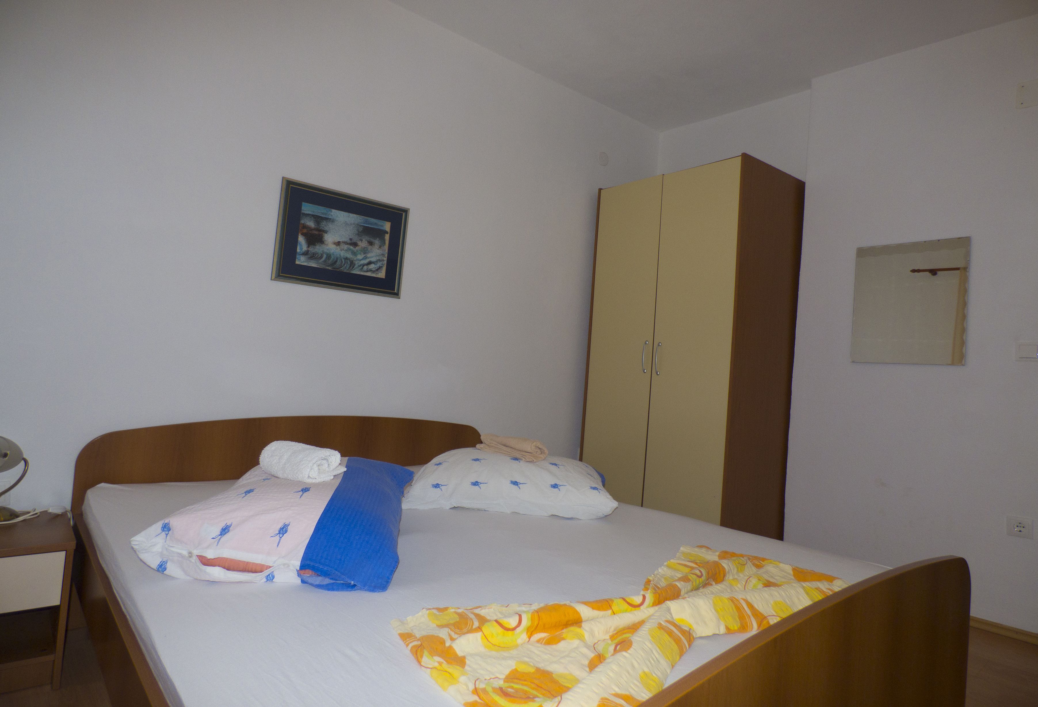 Ein Einzelbett mit Holz Kopfteil, einer Decke mit blau-weißem Muster und einer Decke in Gelb und Orange. Ein Schrank mit brauner Tür und ein gerahmtes Bild an der Wand sind ebenfalls zu sehen.
