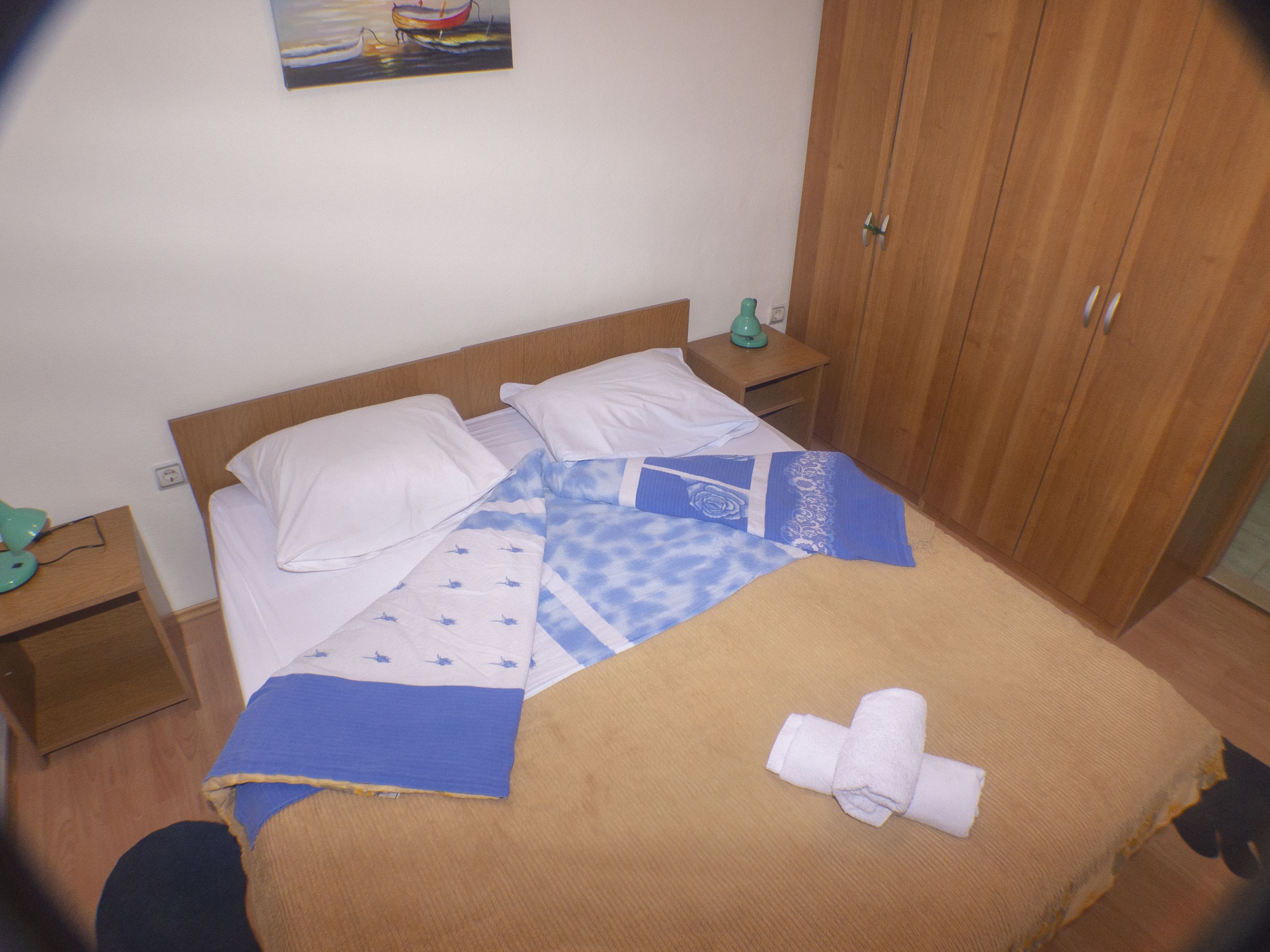 Una cama bien hecha con una manta azul y blanca estampada, un plaid marrón y una toalla blanca enrollada. Se observa un cabecero de madera, una mesita de noche con una lámpara verde y un armario de madera.