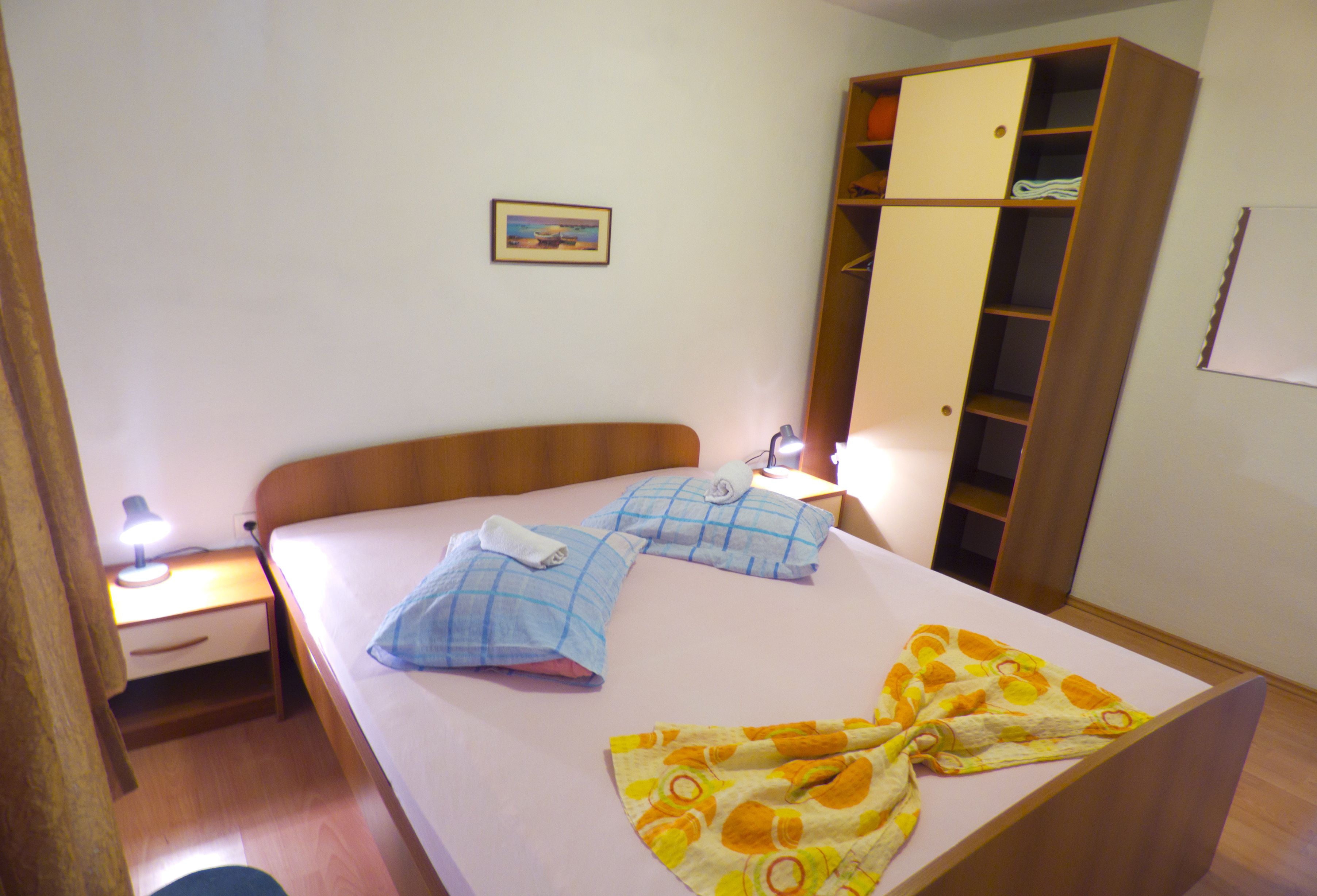 Una cama individual con cabecera de madera, una mesita de noche con lámpara, un armario y un cuadro enmarcado en la pared.
