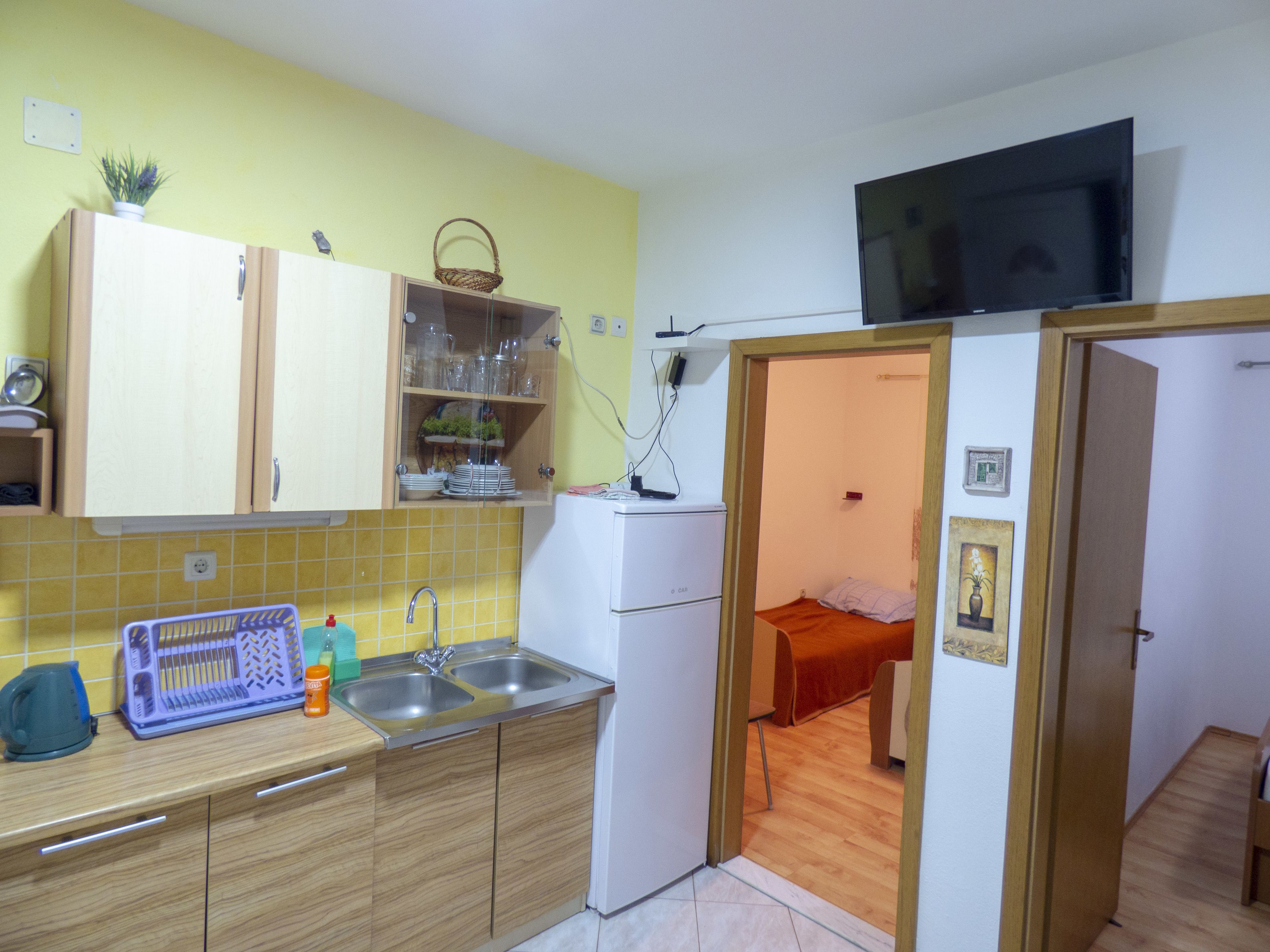 A compact kitchen with wooden cabinets, a sink, and a refrigerator. There is a television mounted above the kitchen counter.