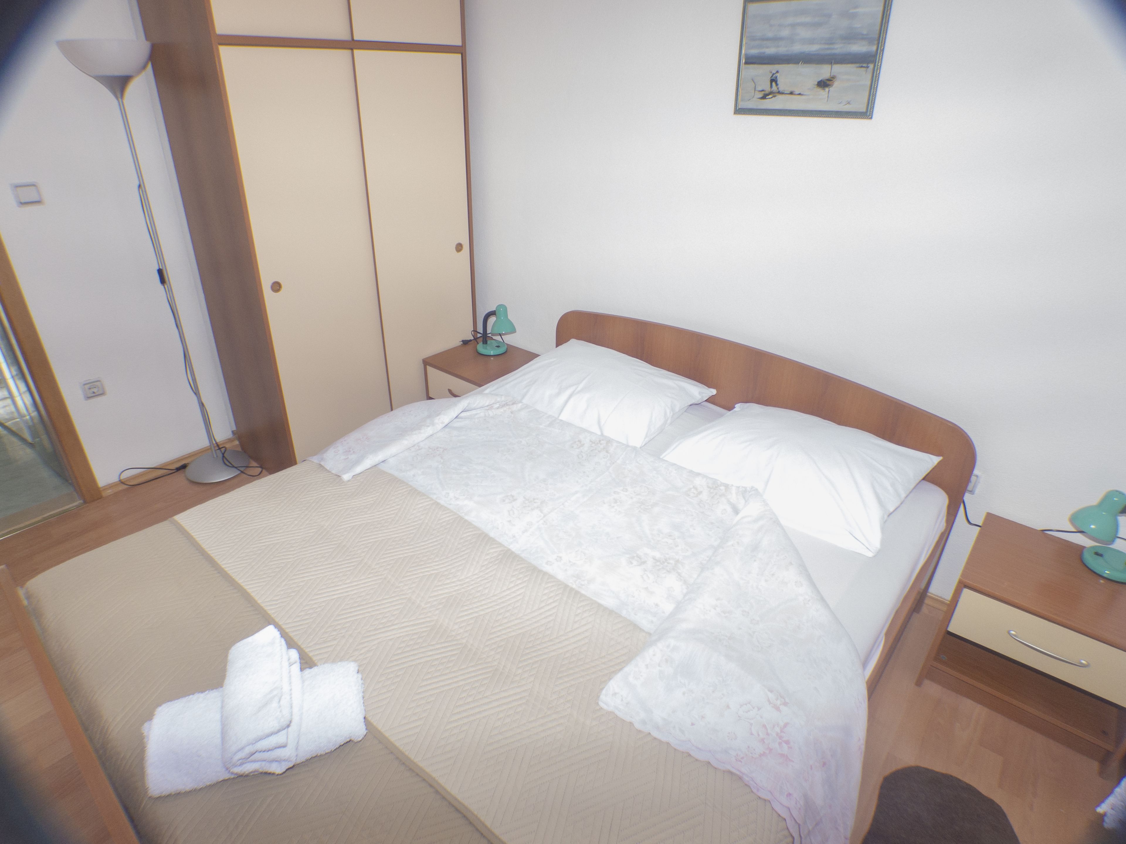 Una cama bien hecha con sábanas blancas, una mesita de noche con lámpara y un pequeño mueble con cajones.