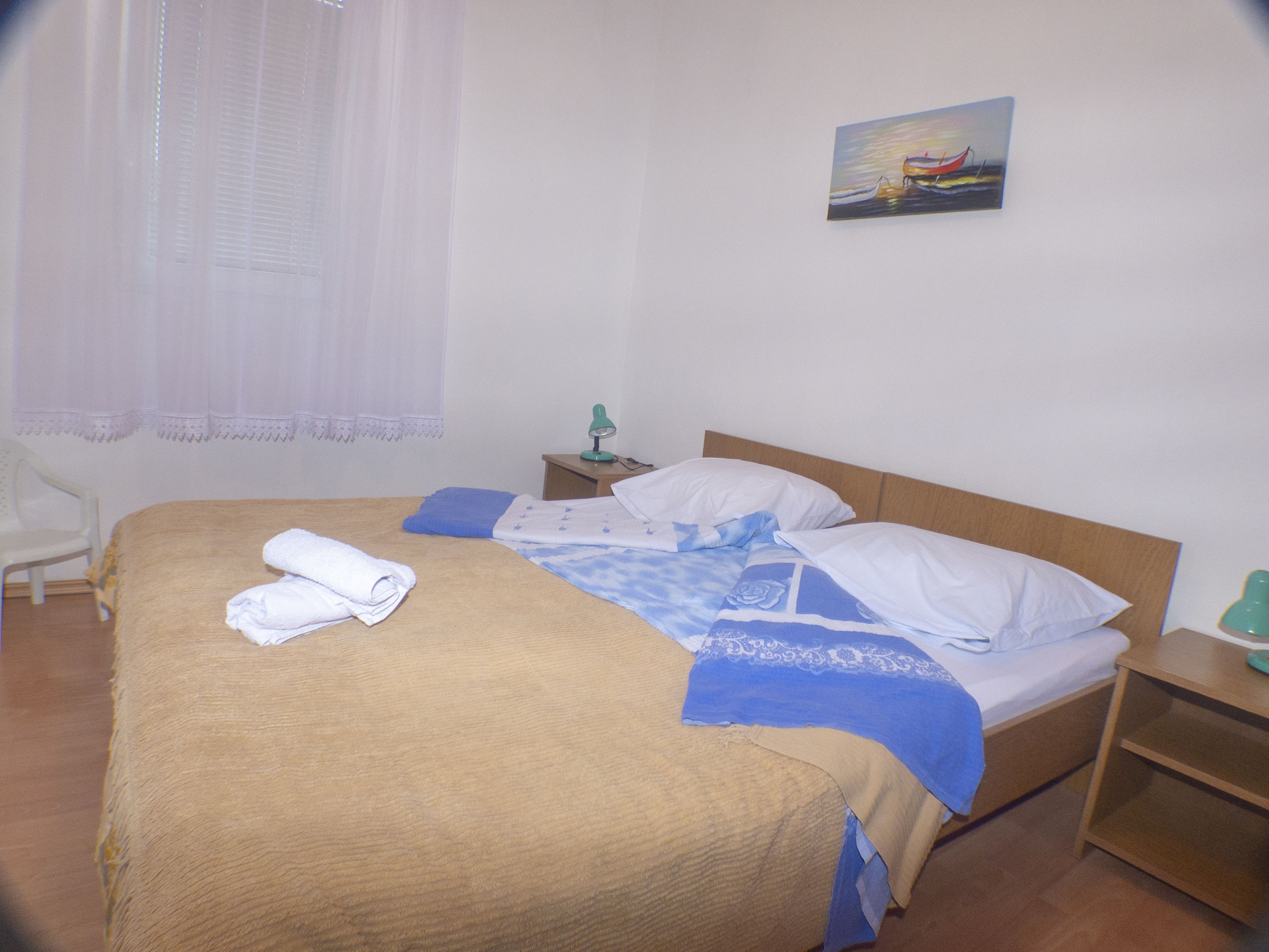 Una cama bien hecha con cubrecama marrón, almohadas azules y blancas, y una manta azul. Hay una mesita de noche con una lámpara, una silla y un cuadro en la pared.