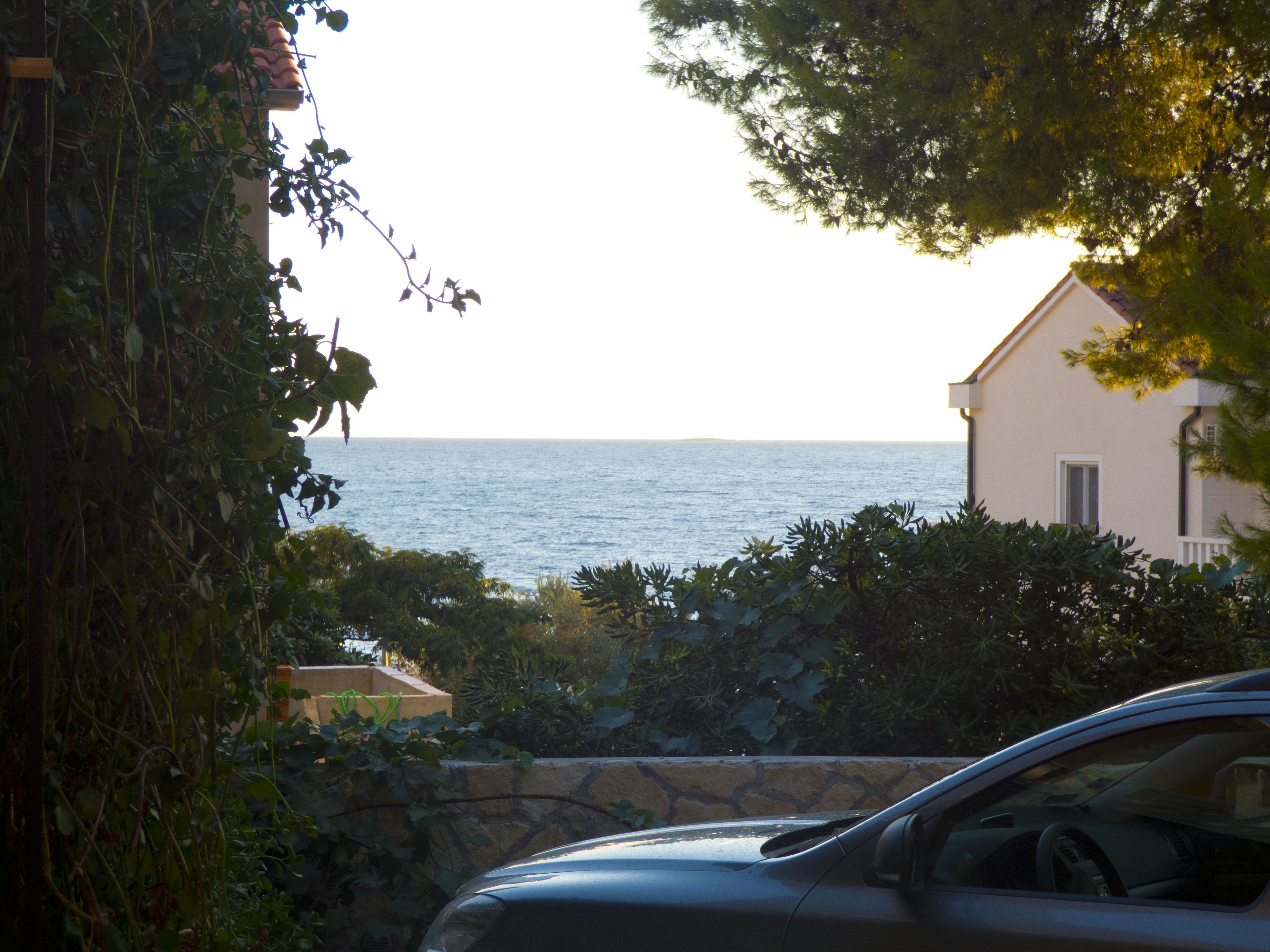 Vista costera con un muro de piedra y un coche al frente, que conduce a un edificio con balcón con vistas al mar.