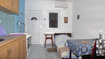 A compact bedroom with a bed, a small table, a chair, a window, and a kitchenette area.