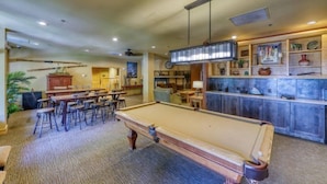 A pool table with surrounding seating, a bar area with shelves of decorative items, and a kitchen area in the background.