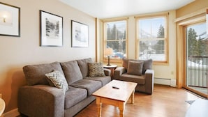 A living room with a sofa, armchair, and coffee table. There are framed pictures on the wall and large windows with blinds.