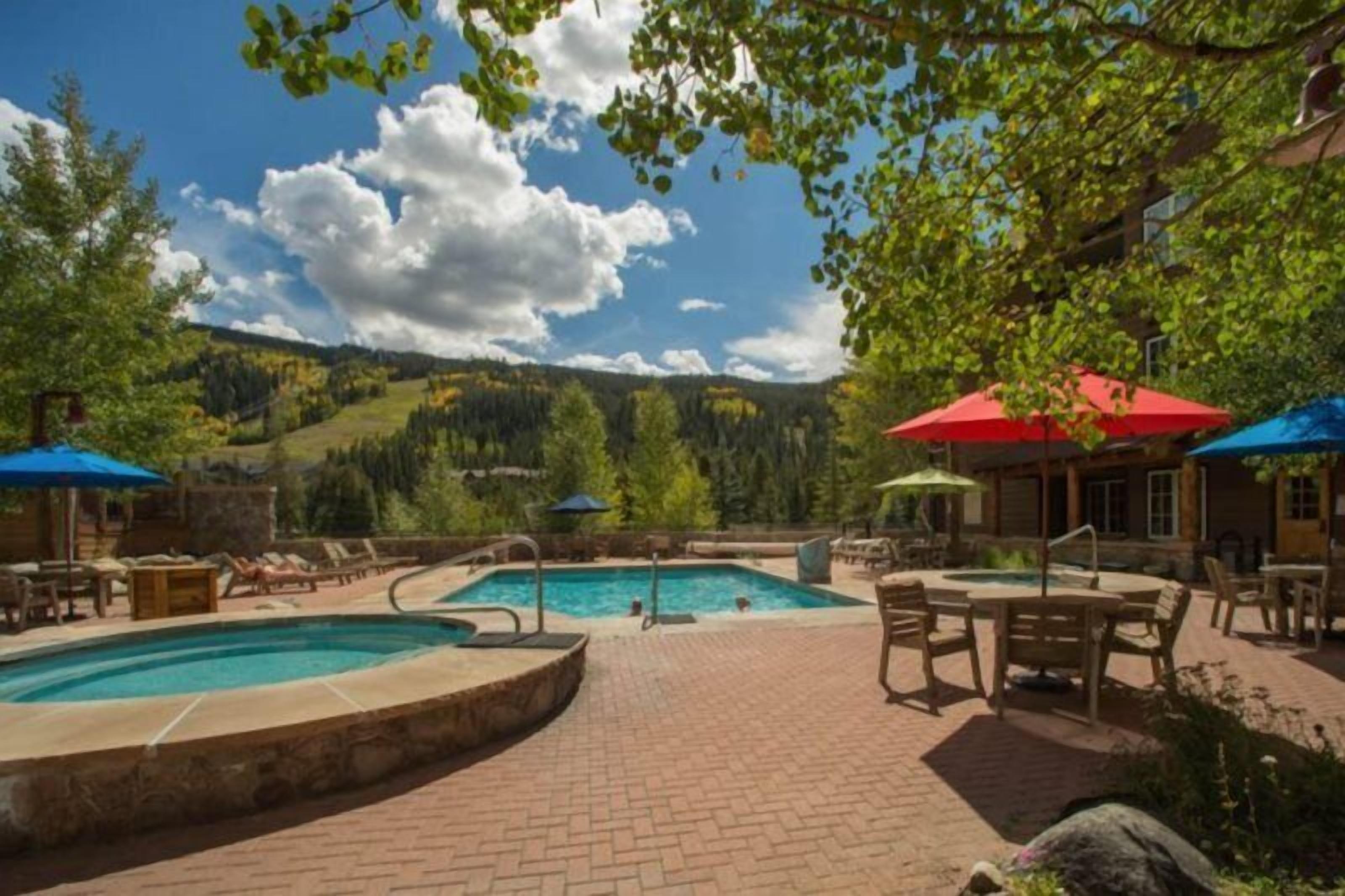 A pool area with lounge chairs and umbrellas, surrounded by a brick patio and trees.