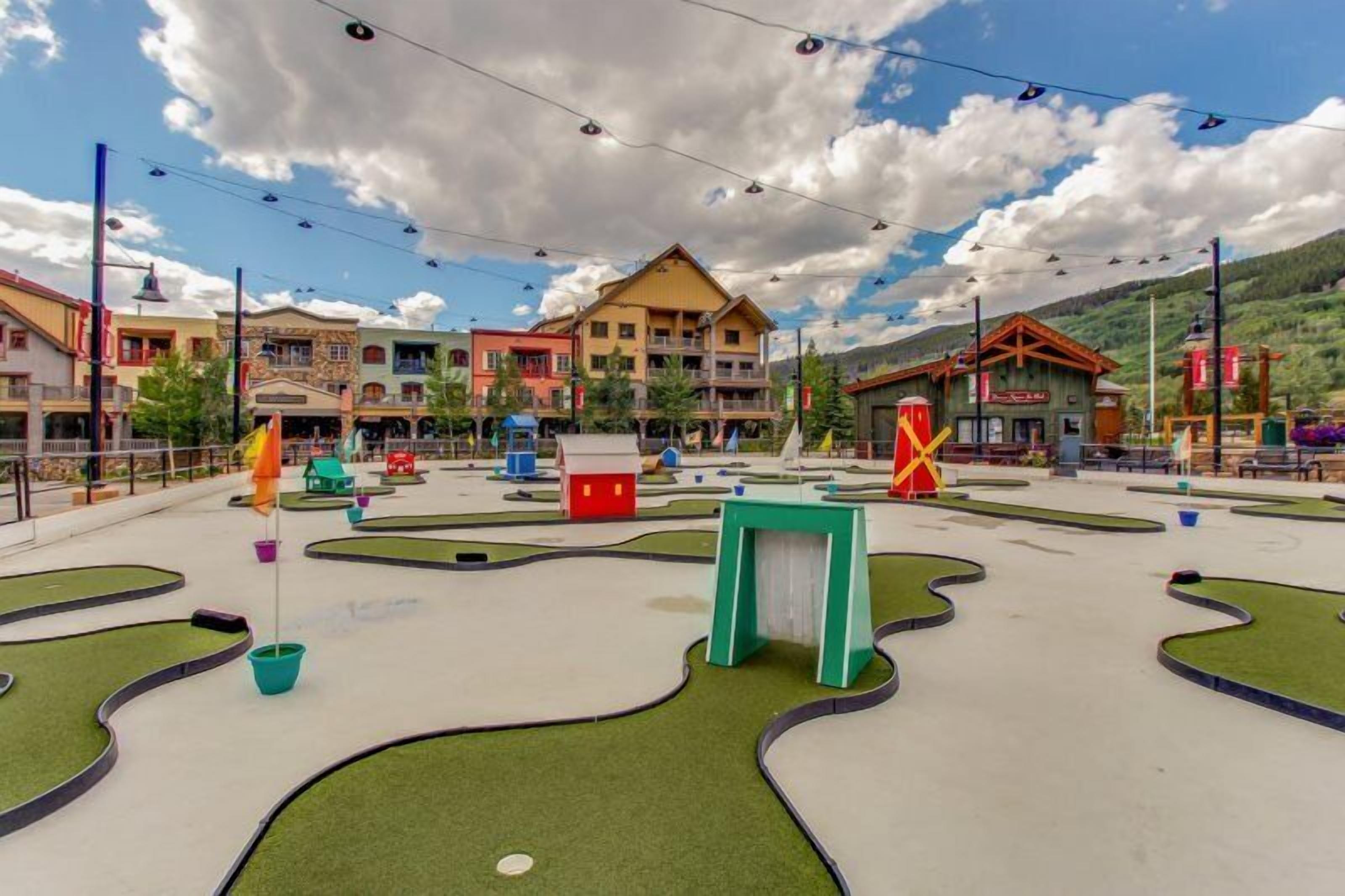 A miniature golf course with a central building, surrounded by colorful buildings and a mountainous backdrop.
