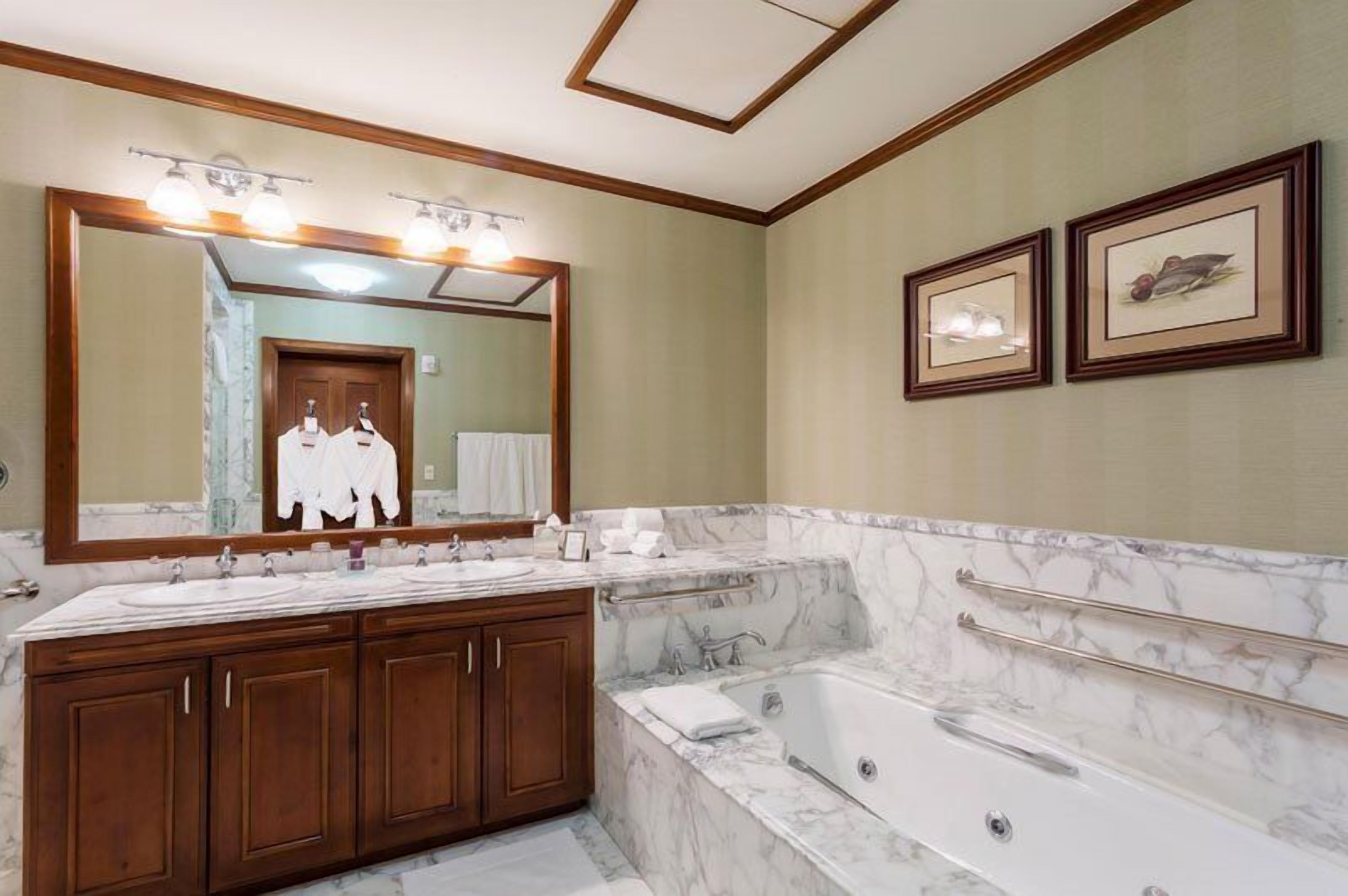 A bathroom with a large mirror, double sinks, a bathtub, and framed artwork on the wall.