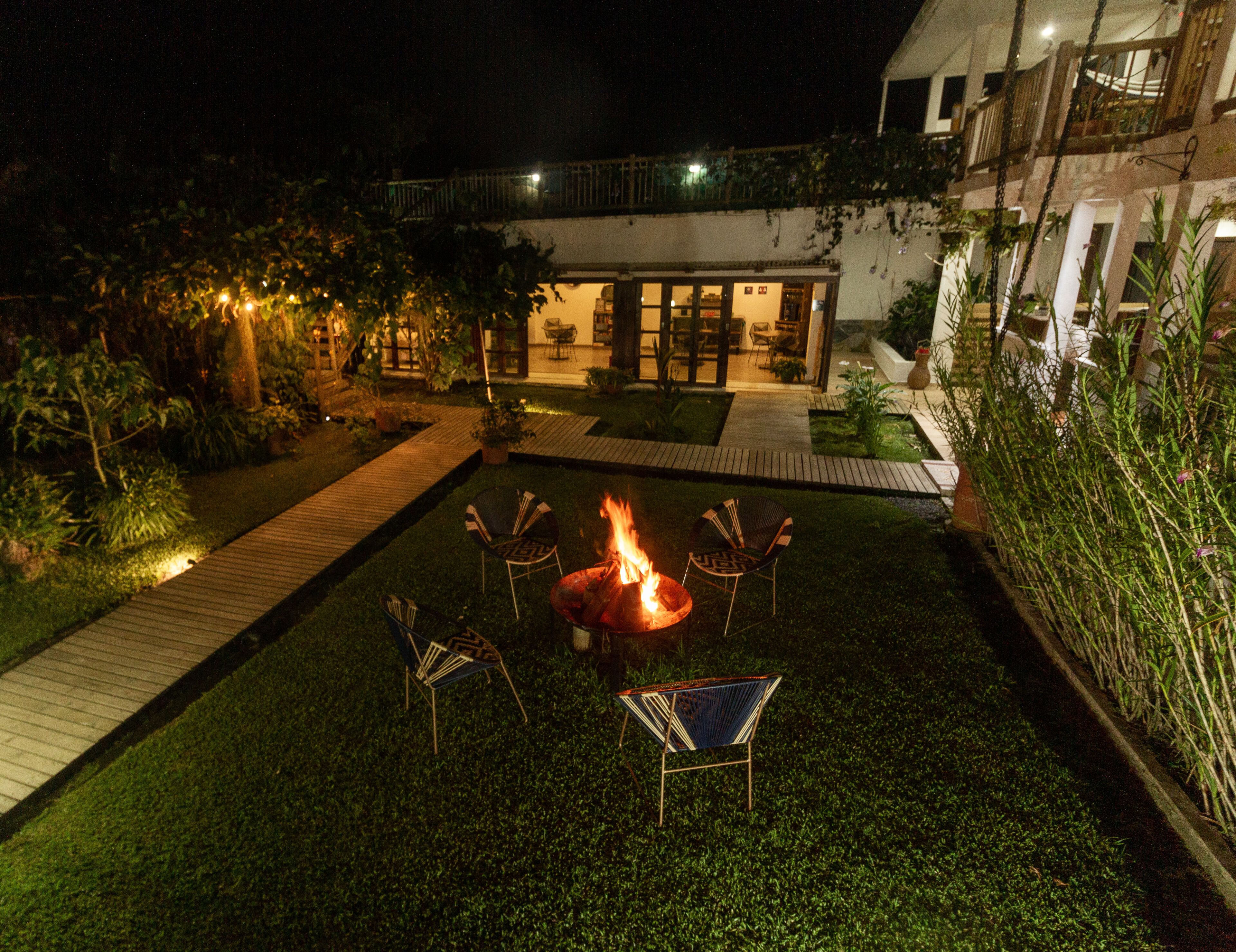 Ein stimmungsvoller Außenbereich mit Sitzgelegenheiten bei Nacht, einem Feuerkorb, umgeben von Stühlen, einer Holzterrasse und einem gepflegten Garten.
