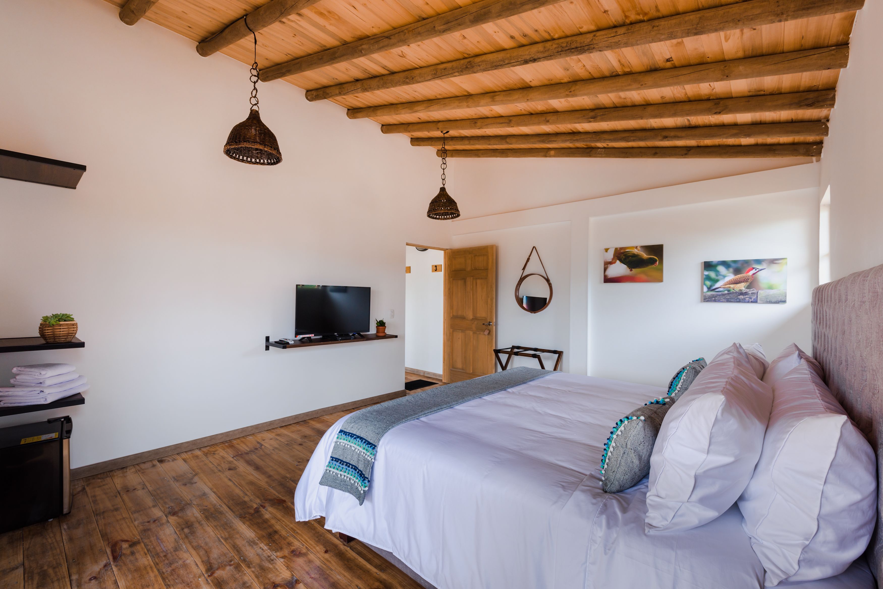 Ein Schlafzimmer mit Holzboden, einem Bett mit weißer Bettwäsche und dekorativen Kissen, einem Fernseher auf einem Regal und einer Tür zu einem anderen Raum.