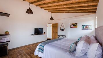 Ein Schlafzimmer mit Holzboden, einem Bett mit weißer Bettwäsche und dekorativen Kissen, einem Fernseher auf einem Regal und einer Tür zu einem anderen Raum.