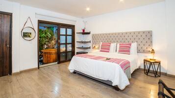 Un dormitorio con cama, mesitas de noche, una mesita auxiliar, suelo de madera, una puerta, una ventana y una estantería con una planta.