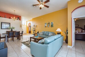 A living room with a blue sofa, a dining area with a table and chairs, and a ceiling fan. - 3BR 2BA Home in Windsor Palms by CV-8168 (Kissimmee)