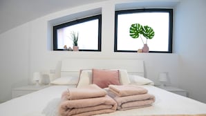 A neatly made bed with pink towels, a white headboard, bedside tables with lamps, and two windows with plants. - Design Central Apartment (Bergamo)