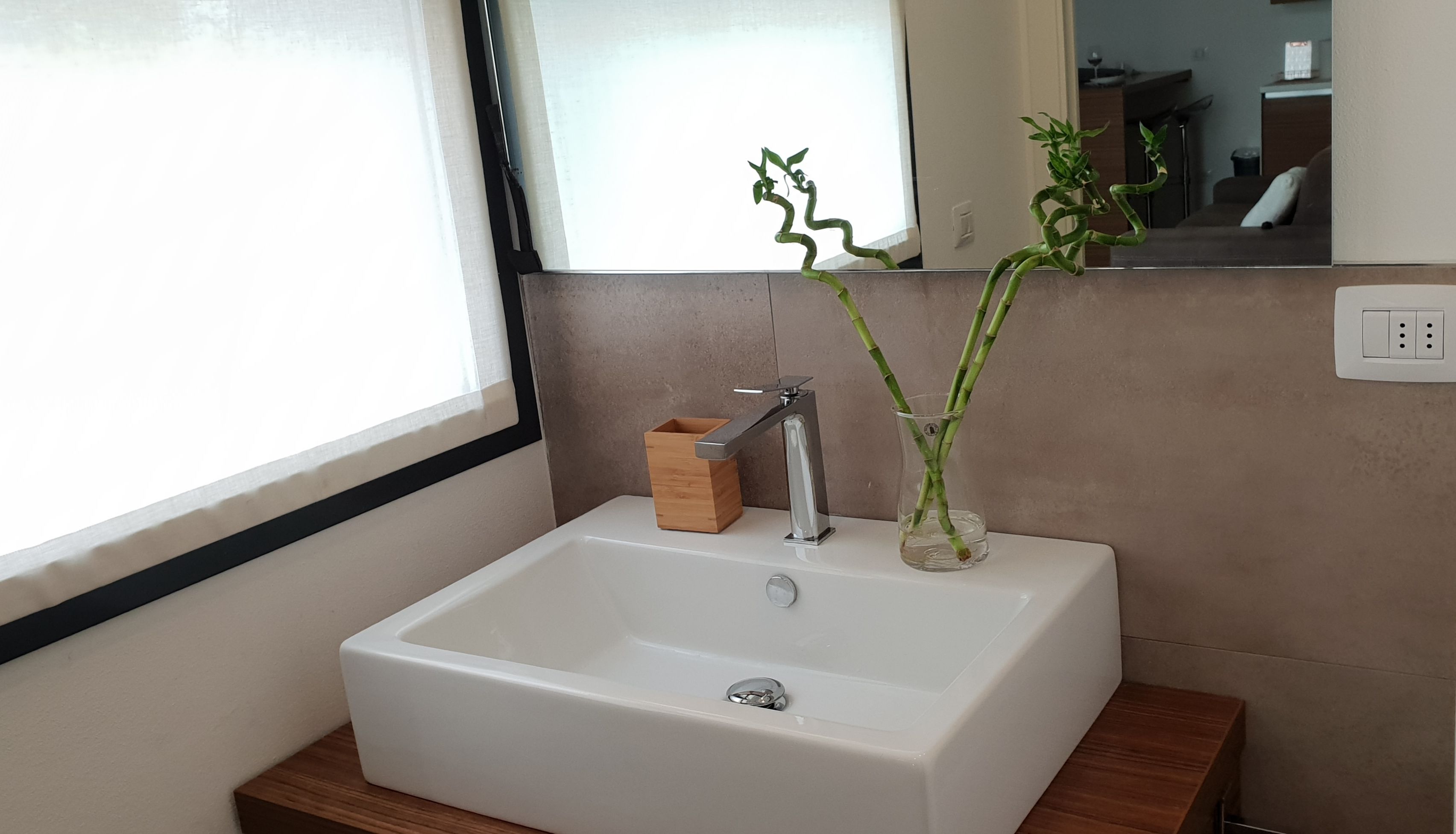 A bathroom with a white sink, a wooden box, and a bamboo plant in a glass vase.