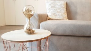 A round wooden table with a copper wire base, a glass hourglass with golden sand, and a grey textured sofa with a white pillow. - Design Central Apartment (Bergamo)
