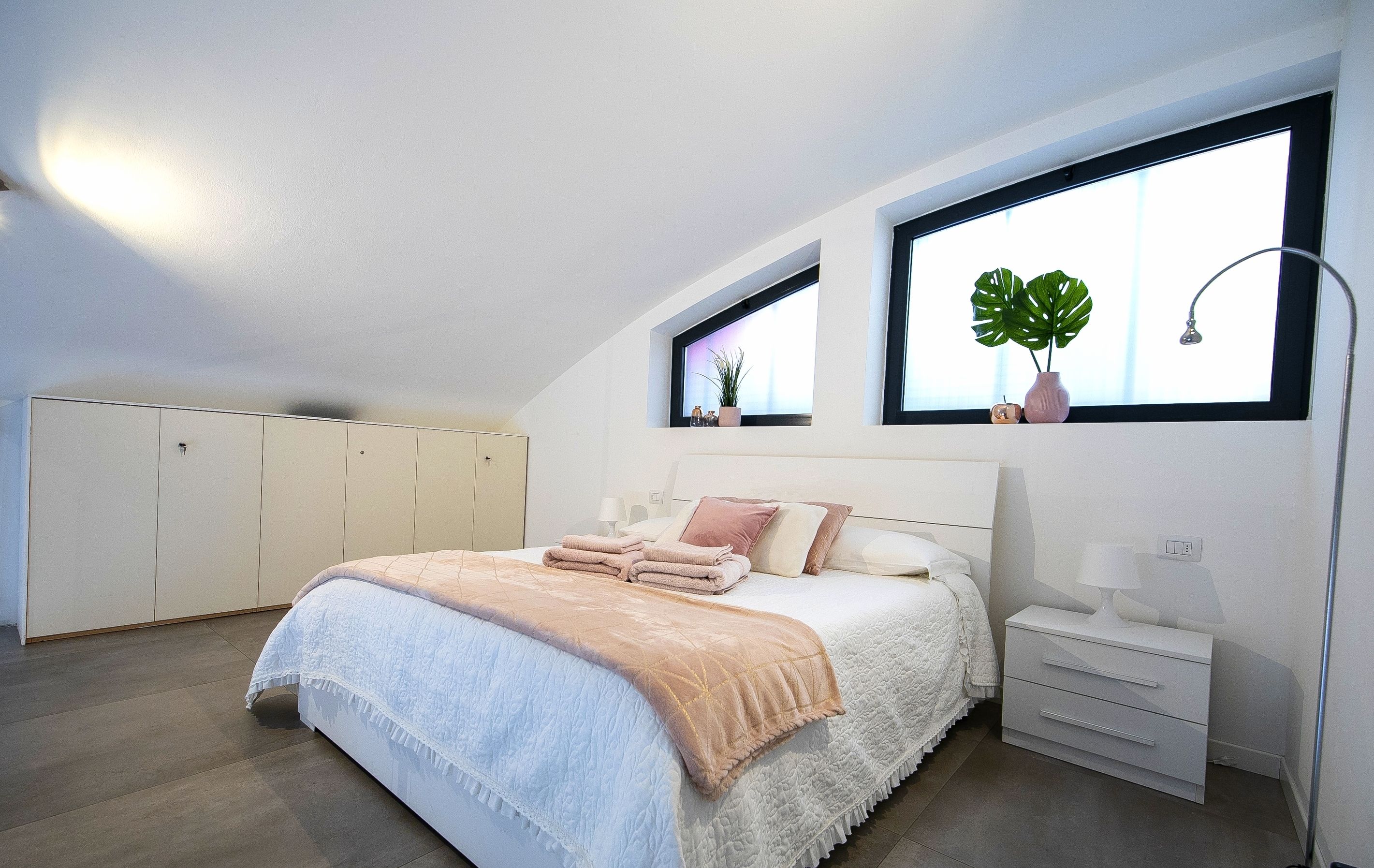 A bedroom with a bed, bedside table, nightstand, and two windows with plants.