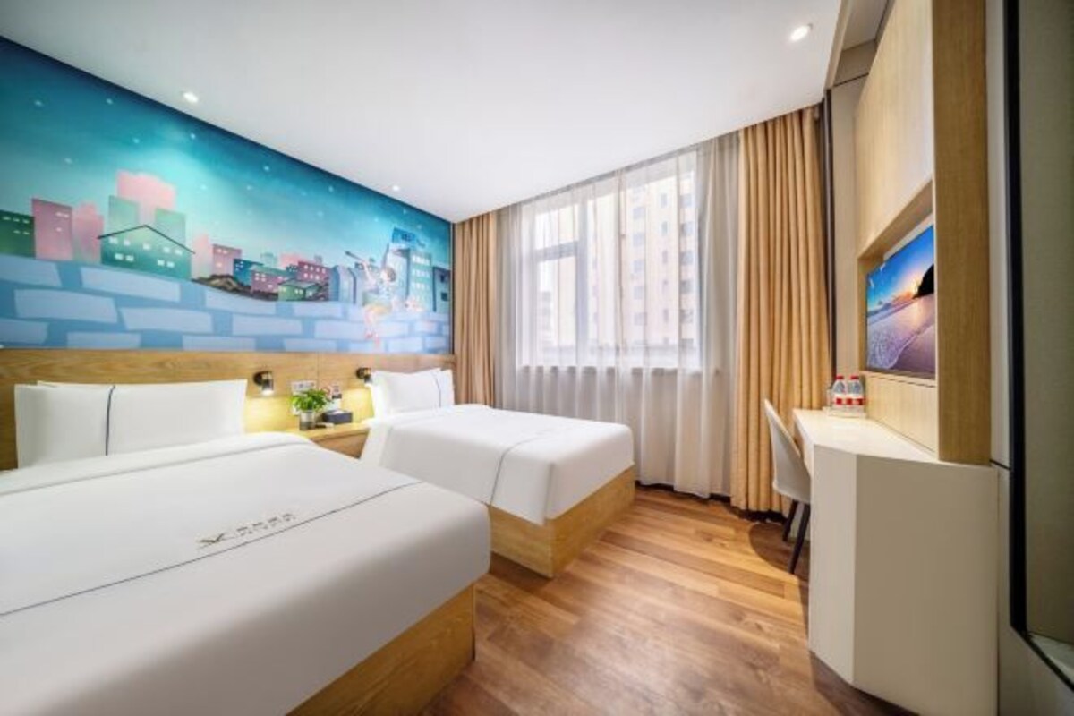 Une chambre d’hôtel avec deux lits, un bureau, une chaise, une fenêtre avec des rideaux et une fresque représentant un paysage urbain sur le mur.