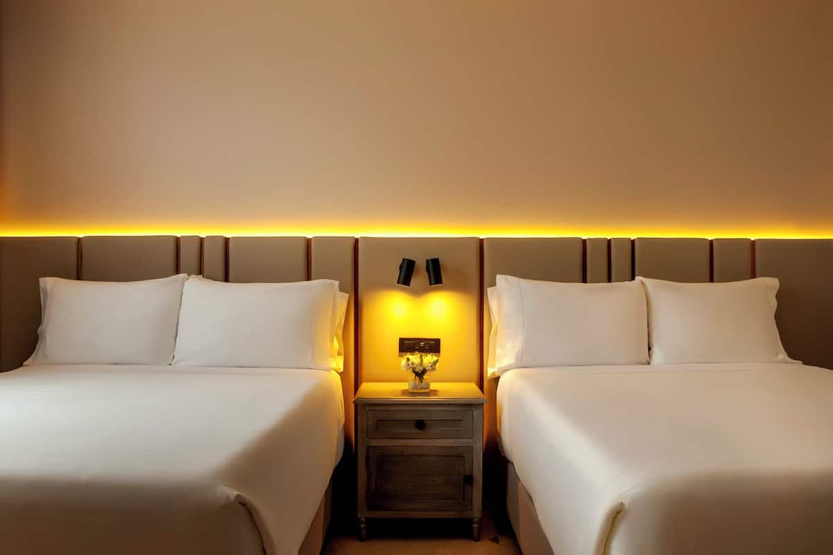 A hotel room with two beds, a nightstand with a lamp, and a wall-mounted light strip.