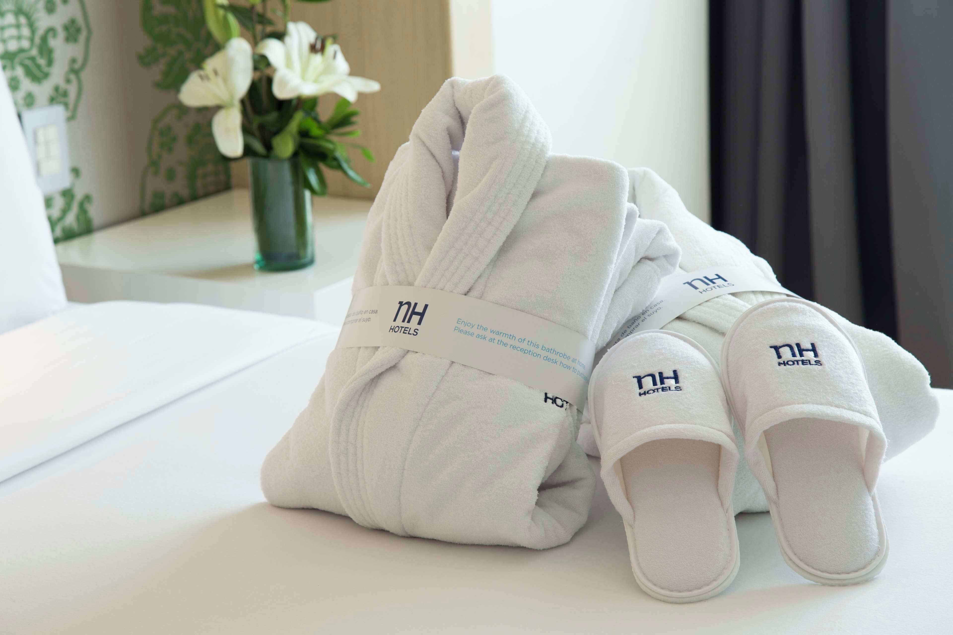 Una toalla blanca doblada con cuidado y zapatillas con el logotipo de "NH Hotels" sobre una superficie blanca y limpia.