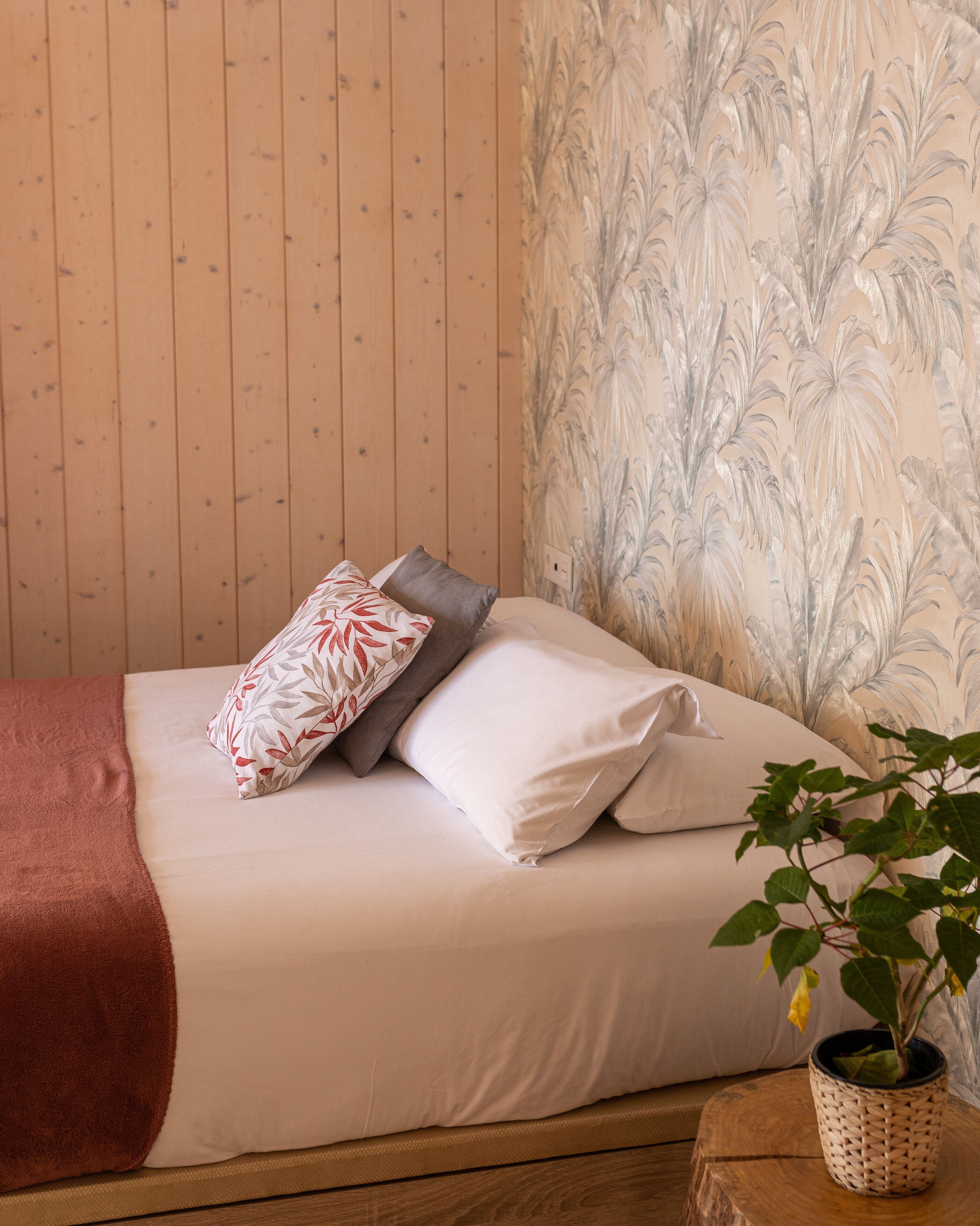 A neatly made bed with a brown blanket, white and patterned pillows, and a potted plant on a wooden nightstand.