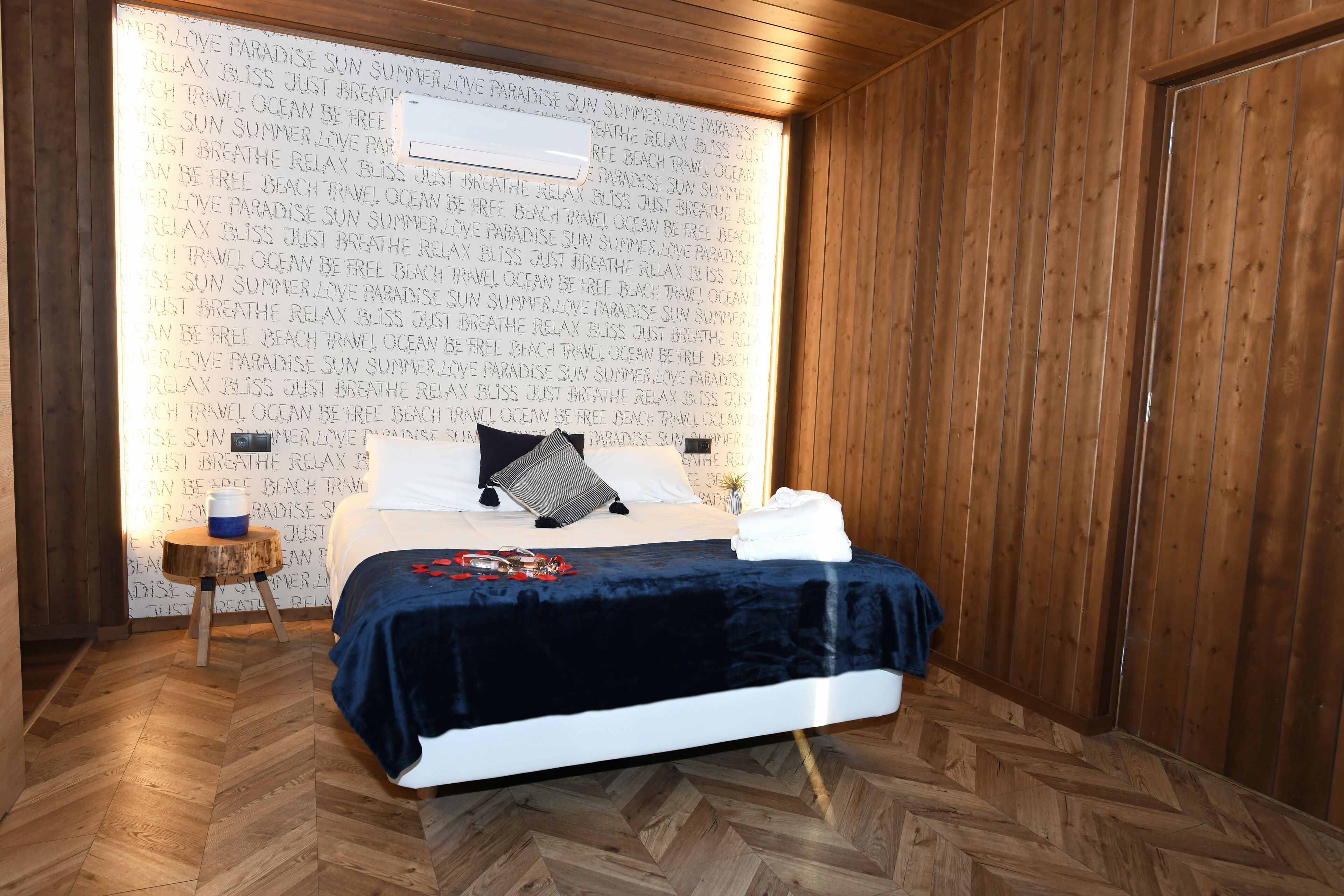 A neatly made bed with a dark blue bedspread, a pillow, and a small wooden stool beside it.