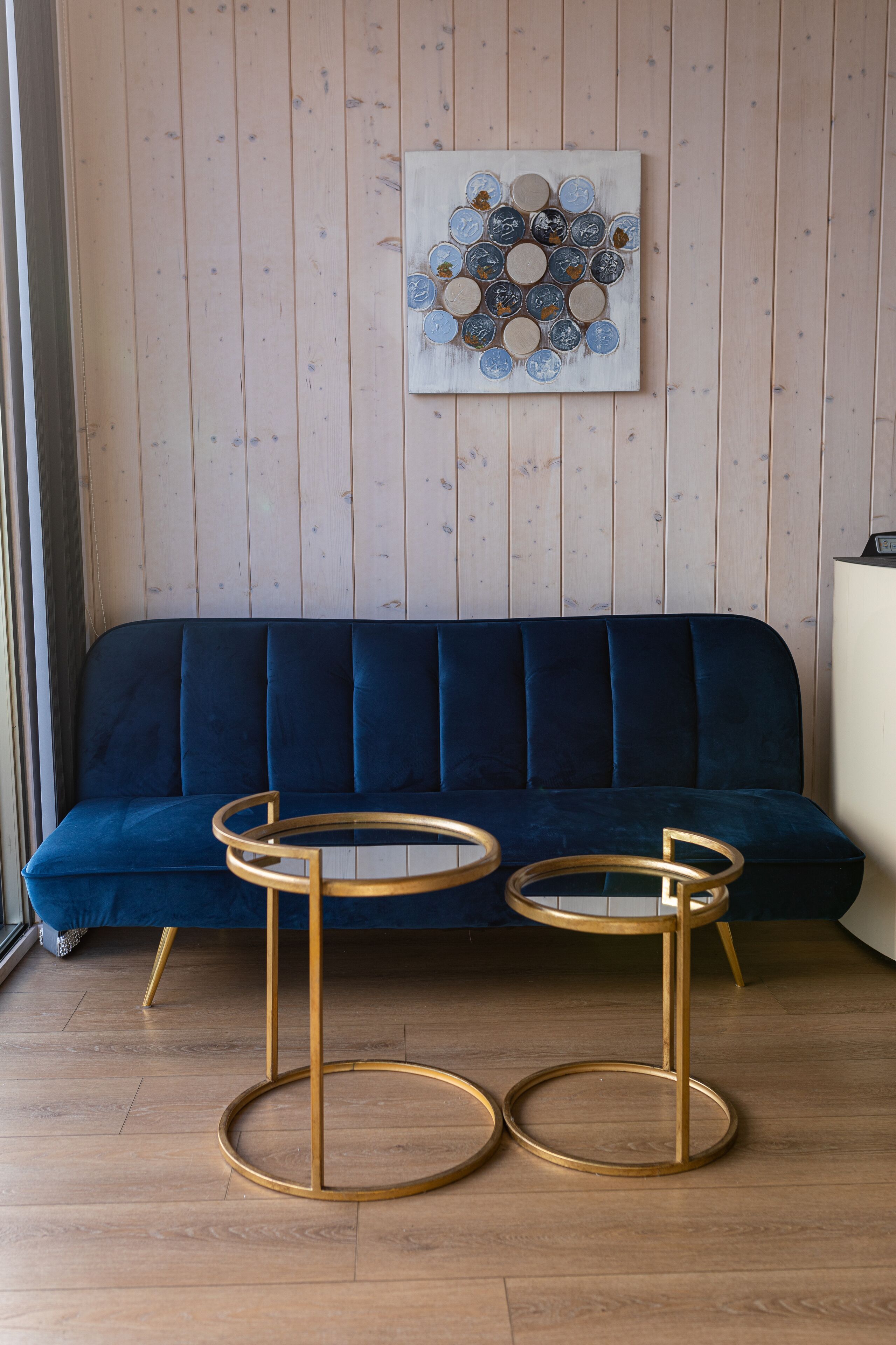 Una habitación con un sofá azul, dos mesas con marcos dorados y una obra de arte colgada en la pared.