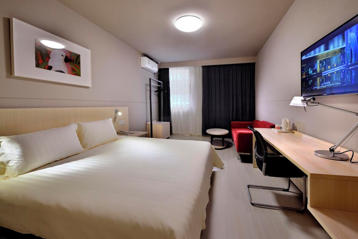 A hotel room with a large bed, a desk with a lamp, a television, and a red chair.