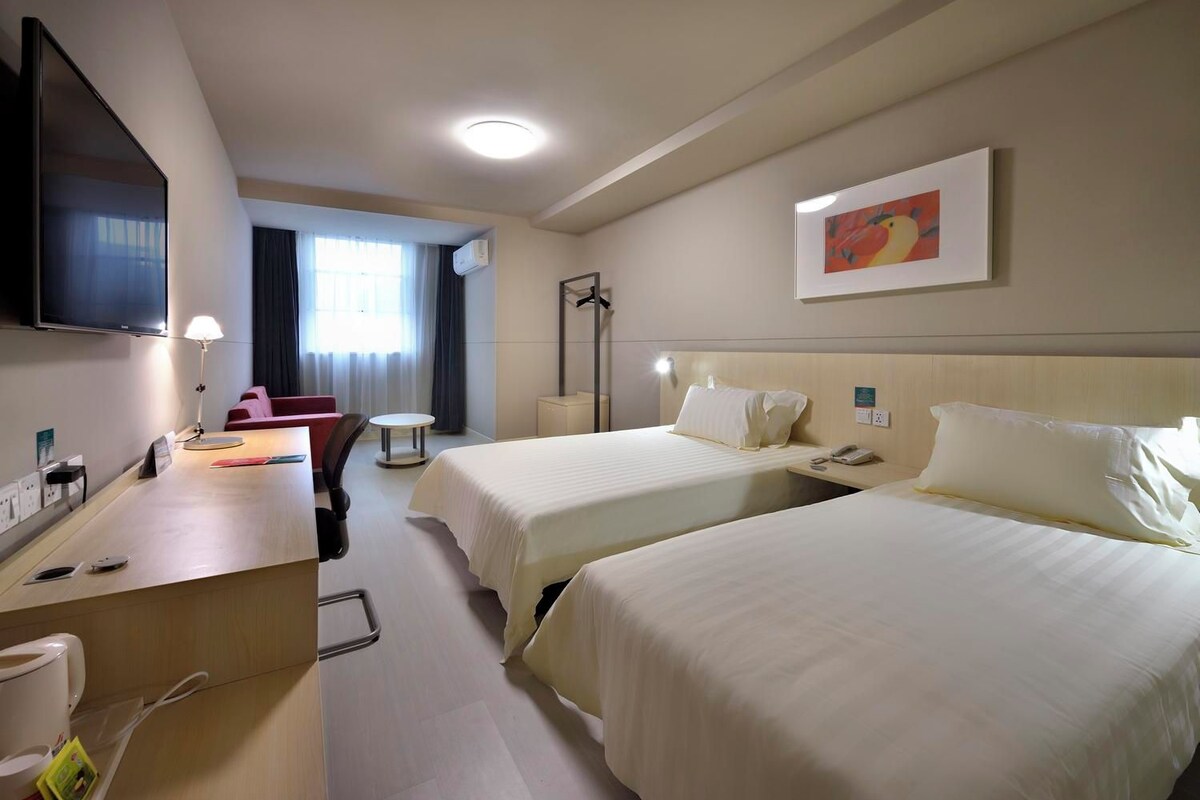 A hotel room with two beds, a desk with a lamp, a TV, and a painting on the wall.