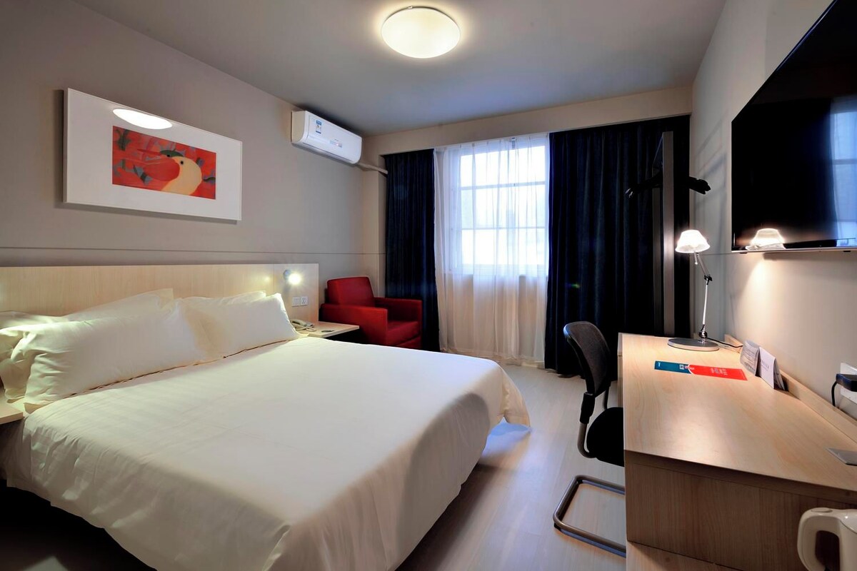 A hotel room with a large bed, a desk with a lamp, a chair, a TV, and a window with curtains.