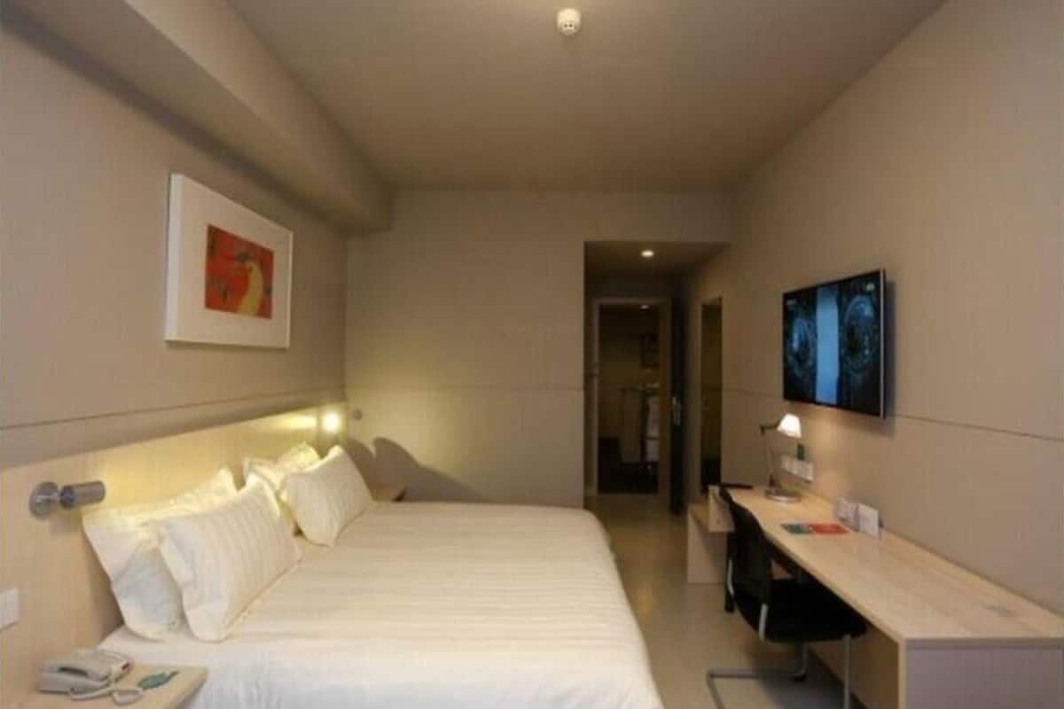 A hotel room with a bed, bedside table, lamp, desk, chair, and a television on the wall.