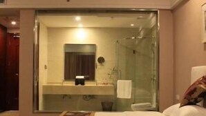 A hotel room with a glass-enclosed shower, a small bathroom with a mirror, and two beds with a patterned bedspread. - Airport Hotel Hefei (Hefei)