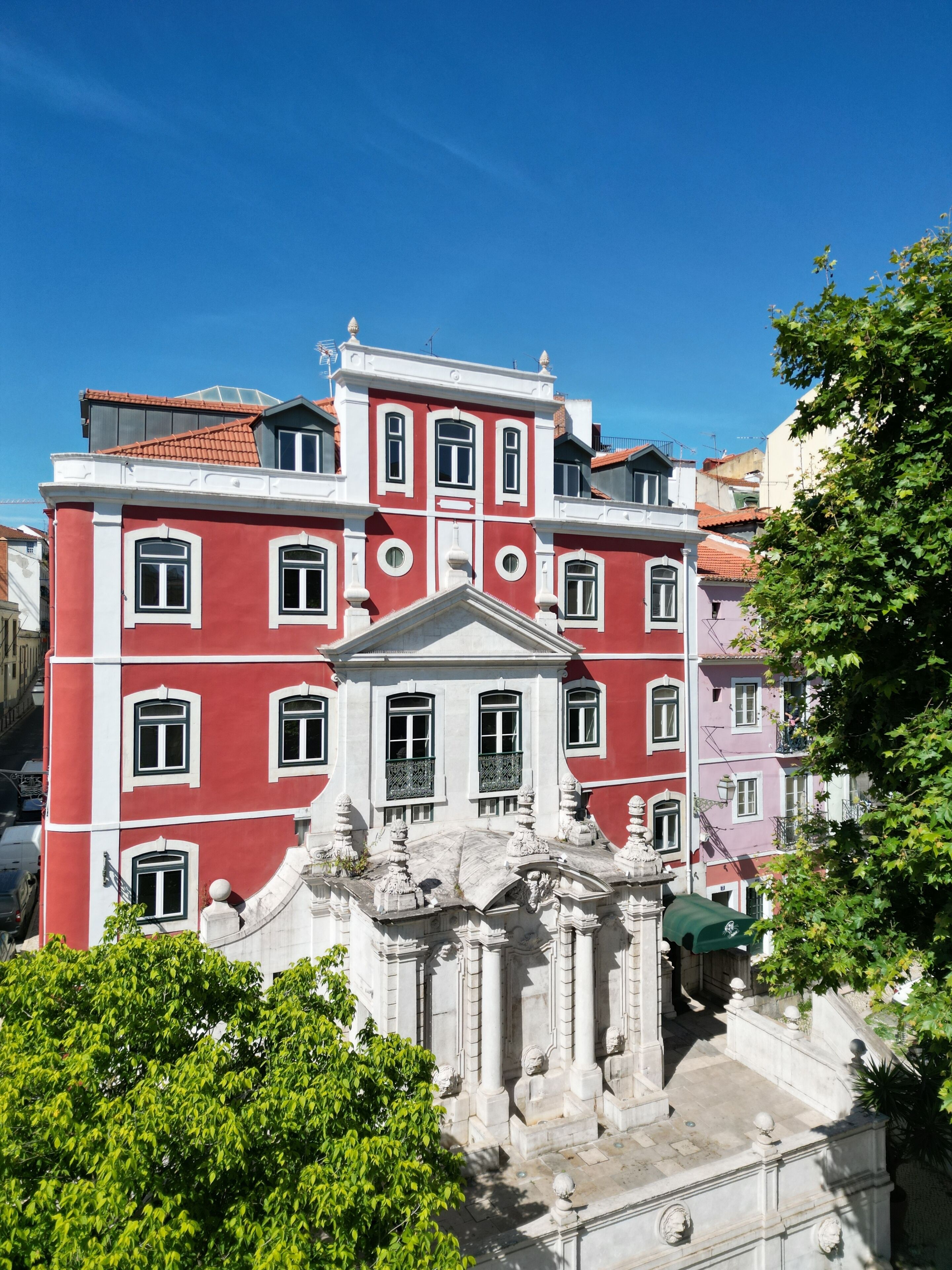 A multi-story building with classical architecture, featuring a central entrance with columns and a pediment, surrounded by lush greenery.