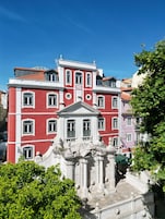 Un edificio de varios pisos con arquitectura clásica, que presenta una entrada central con columnas y un frontón, rodeado de abundante vegetación.