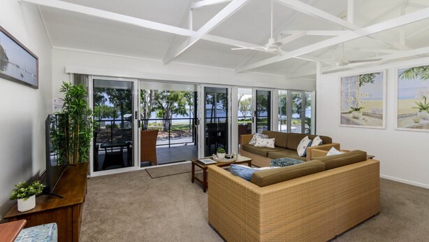 Un vaste salon comprenant un grand canapé, une table basse et un coin repas avec une table et des chaises. De grandes portes-fenêtres coulissantes donnent sur une terrasse extérieure offrant une vue sur la mer.