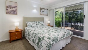 Une chambre à coucher comprenant un lit, deux tables de chevet, un ventilateur de plafond et un balcon avec vue sur la verdure.