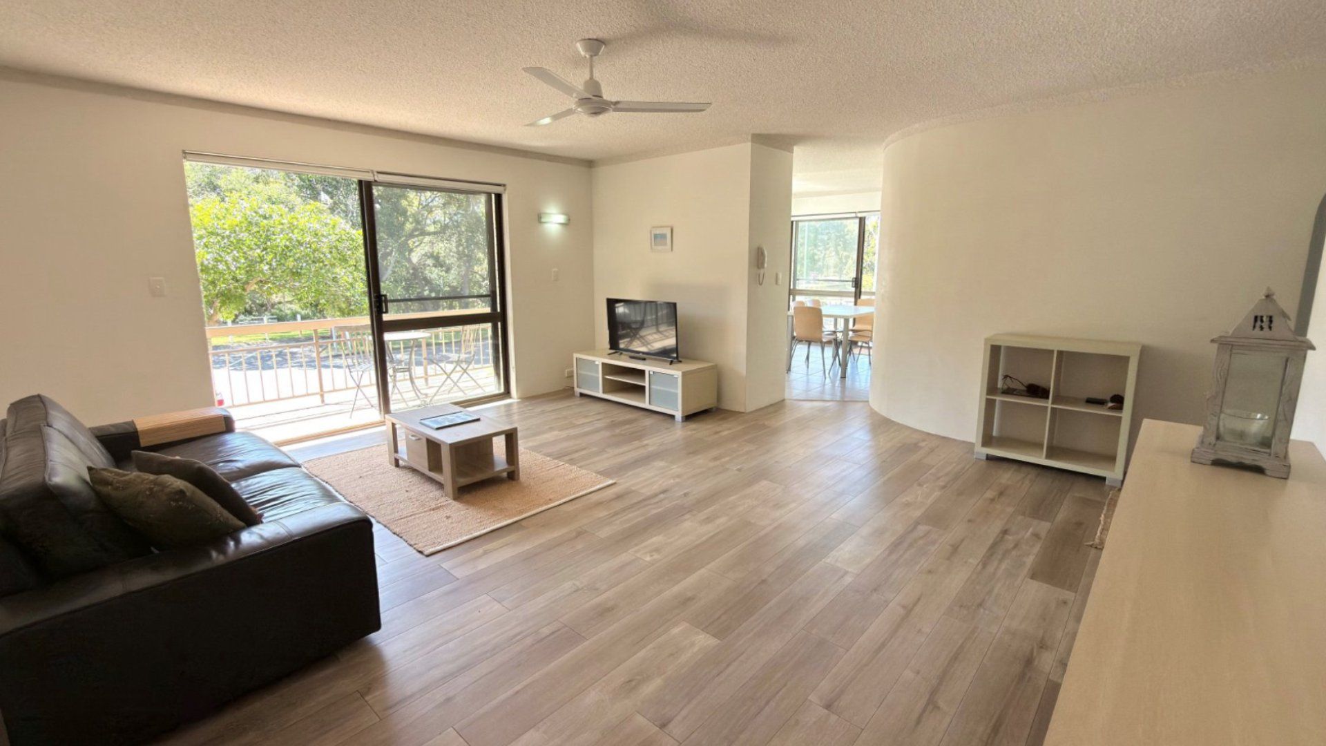 A spacious living room with a dark leather sofa, a TV on a white stand, and a balcony with a view of trees.