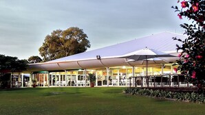 A covered outdoor seating area with tables and chairs, surrounded by a grassy lawn and a large tree.